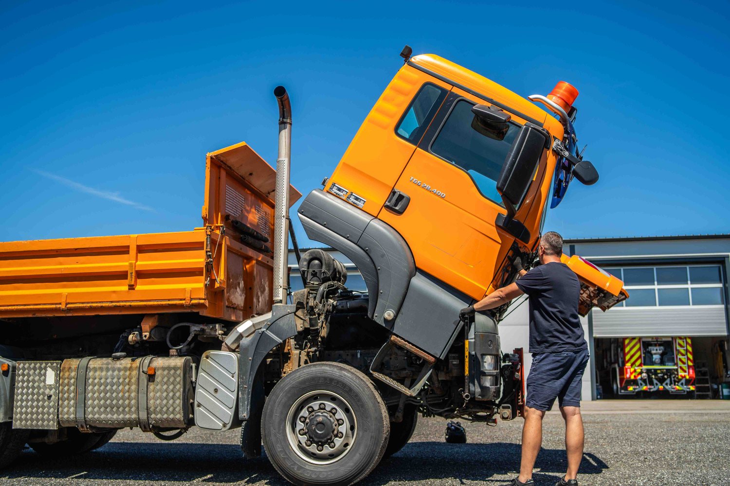 Felbillentett vezetőfülkéjű narancssárga billencs a telephelyen, szakember a motortérnél