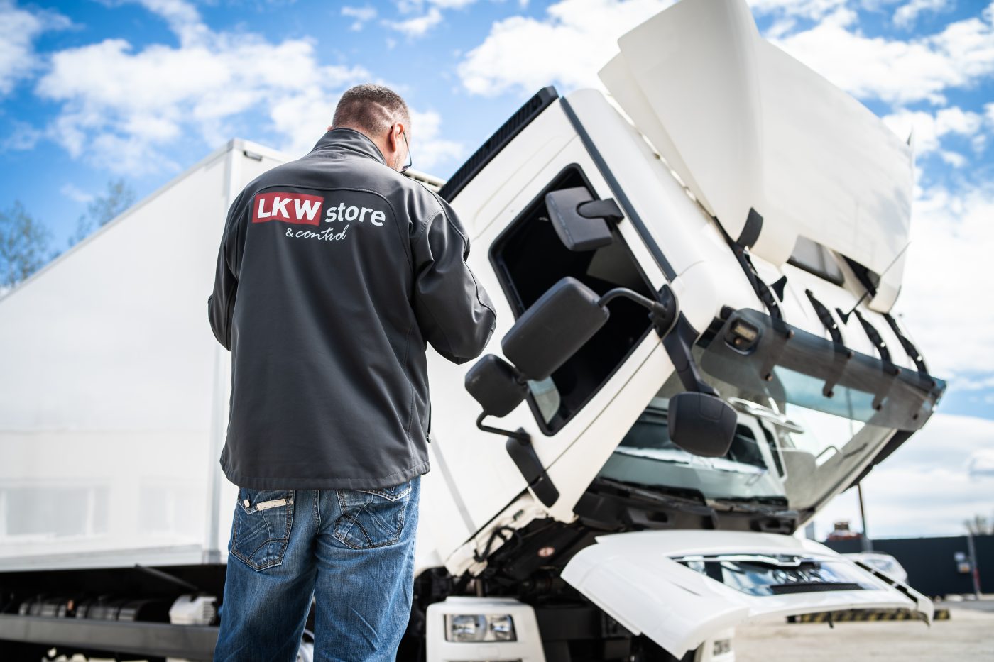 LKW Store feliratú kabátot viselő szakember egy felbillentett fülkéjű fehér teherautó előtt